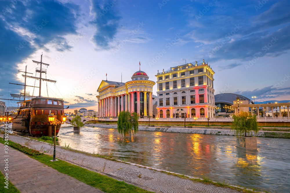 Beautiful sunset in Skopje with light reflection on Vardar river. The archaeological Museum of Macedonia and the Bridge of Civilizations at sunset. Old ship in the river.
