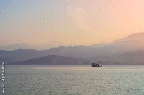 Fishing vessel sails on Black sea at dawn with view of misty mountains on other shore with shining sun in Batumi, Georgia