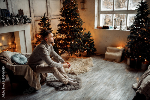 Charming man sitting in cozy chair in living room near the fireplace and Christmas tree, thinking about presents for family on winter holidays, new year celebration concept