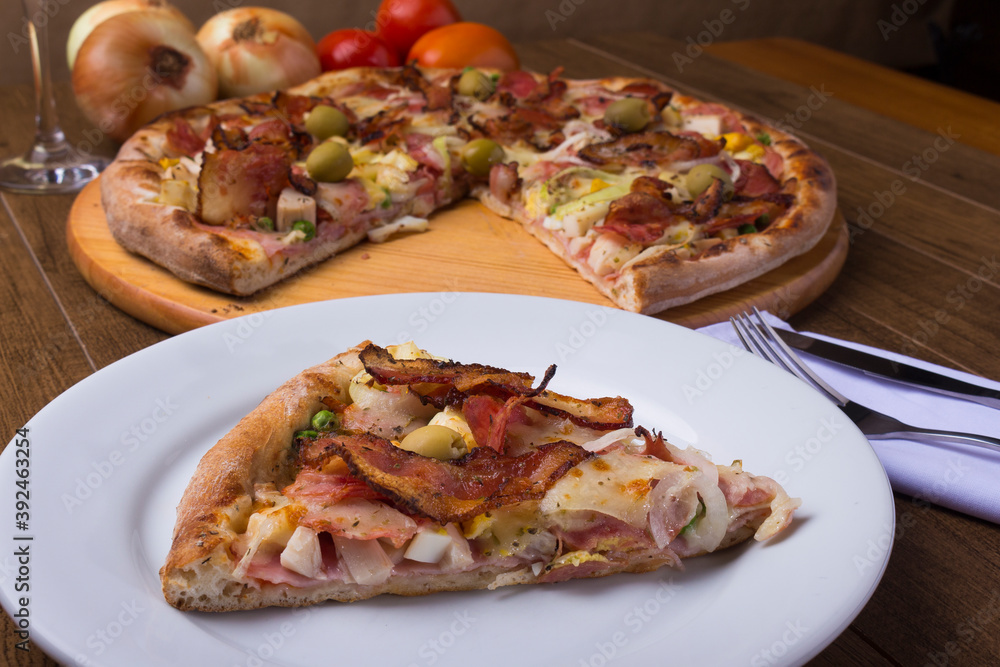 Portuguese Pizza slice made with ham, pea egg, heart of palm, pepperoni, onion and mozzarella and bacon. Served on the plate with sliced pizza in the background. Close-up photo for pizzerias. Portuguese Pizza slice made with ham, pea egg, heart of palm, pepperoni, onion and mozzarella and bacon. Served on the plate with sliced pizza in the background. Close-up photo for pizzerias.