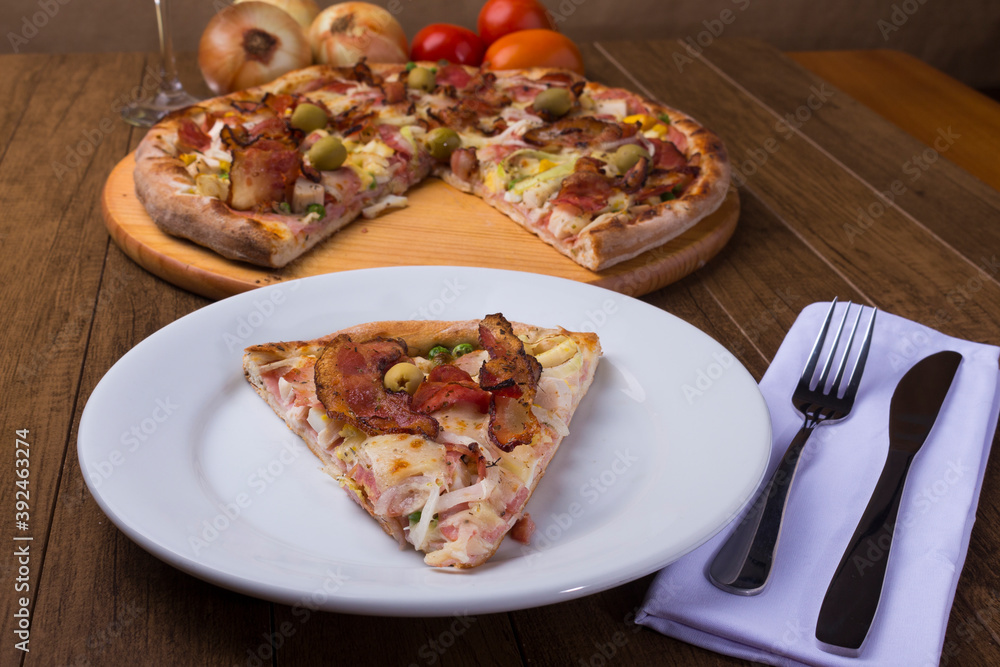 Portuguese Pizza slice made with ham, pea egg, heart of palm, pepperoni, onion and mozzarella and bacon. Served on the plate with sliced pizza in the background. Portuguese Pizza slice made with ham, pea egg, heart of palm, pepperoni, onion and mozzarella and bacon. Served on the plate with sliced pizza in the background.