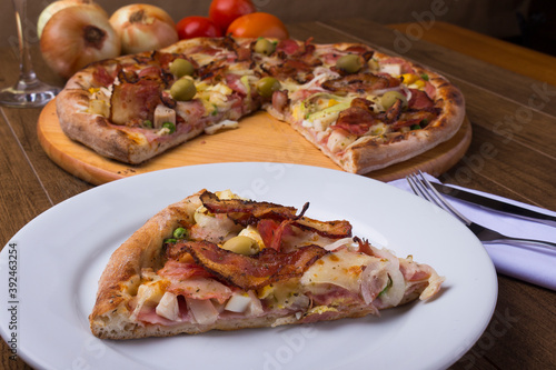 Portuguese Pizza slice made with ham, pea egg, heart of palm, pepperoni, onion and mozzarella and bacon. Served on the plate with sliced ​​pizza in the background. Close-up photo for pizzerias.
