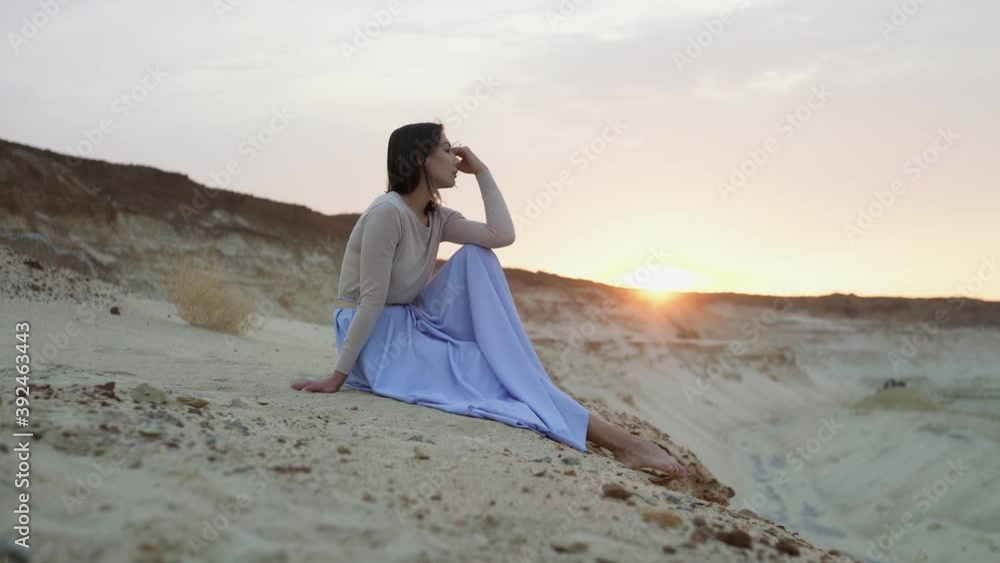 Beautiful young woman in blue skirt sits on top of sand hill with sunset on background Beautiful young woman in blue skirt sits on top of sand hill with sunset on background