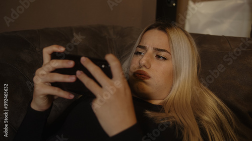 Young blond beautiful woman stretching on the sofa is playing game with smartphone and feeling sad
