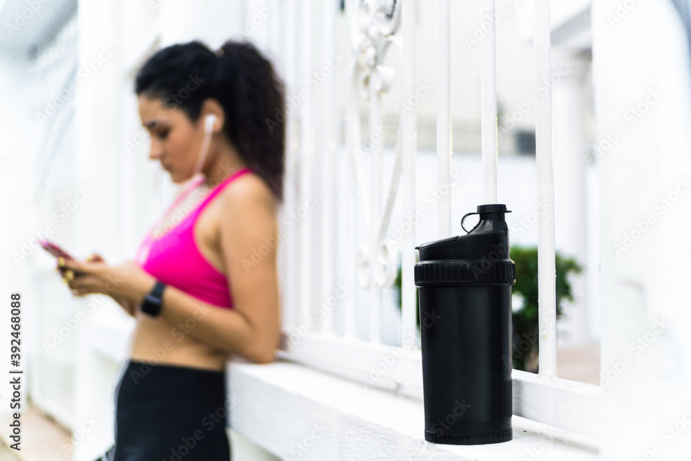 selective focus of thermo of drink and hispanic young woman using mobile phone in outdoors after running. healthy concept. selective focus of thermo of drink and hispanic young woman using mobile phone in outdoors after running. healthy concept.
