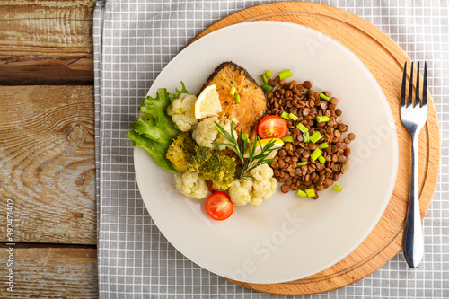 Baked fish with broccoli and lentils in a white plate with rosemary and lemon on a napkin and a round stand next to a fork.