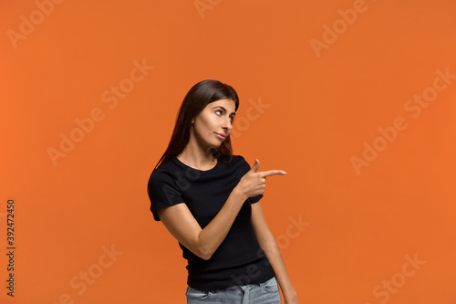 Look at this. Smiling caucasian woman in black t-shirt looking and points to side shows good advertisement, advises to pay attention. Isolated over an orange background