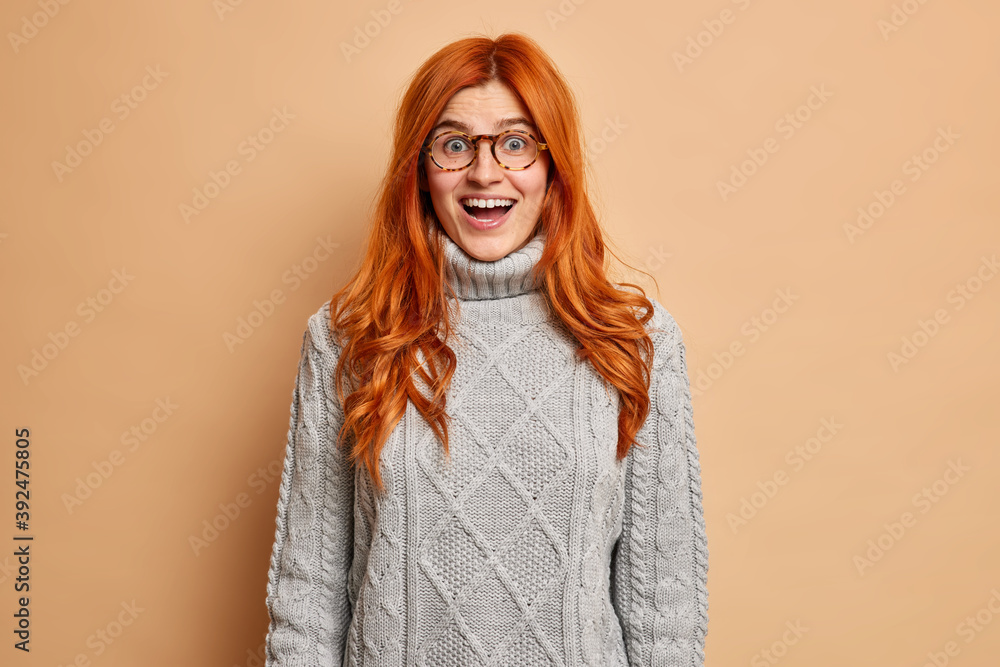 Happy surprised redhead woman looks with opened mouth at camera cannot believe in her sudden success dressed in knitted sweater isolated over brown background. Emotions and reaction concept.
