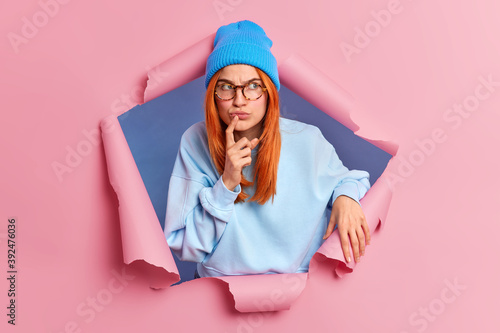 Serious pensive redhead young woman looks away with thoughtful concentrated face expression keeps index finger near lips wears blue clothing poses through pink torn background. Deep thoughts © Wayhome Studio