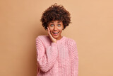 Surprised happy young woman with Afro hair looks with excitement and broad smile keeps hand on face dressed in casual knitted jumper poses against beige studio background. Human reactions concept