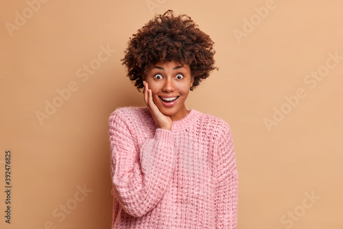 Surprised happy young woman with Afro hair looks with excitement and broad smile keeps hand on face dressed in casual knitted jumper poses against beige studio background. Human reactions concept
