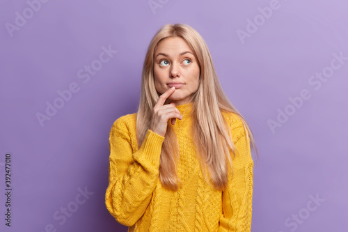 Blonde attractive young European woman keeps finger on lips looks with pensive expression above makes important decision builds plans in mind wears yellow sweater isolated over purple background