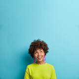 Pleased dark skinned woman looks happily above smiles pleasantly notices something pleasant has curly hair dressed in casual t shirt isolated over blue background. Vertical shot. Copy space for promo