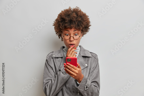 Emotive curly haired ethnic woman stares impressed at smartphone display has bugged eyes reads shocking news or comment stares with disbelief wears round glasses and grey jacket poses indoor.