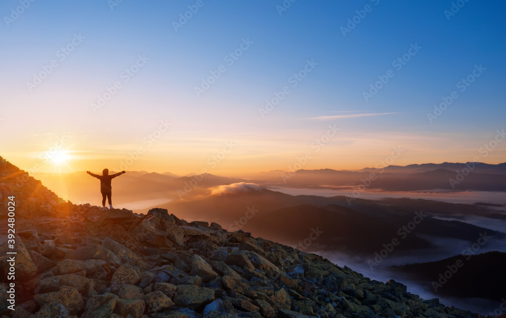 Girl on a mountain top with outstretched arms. Dramatic landscape of a lone hiker looking at mountains in fog at sunrise.