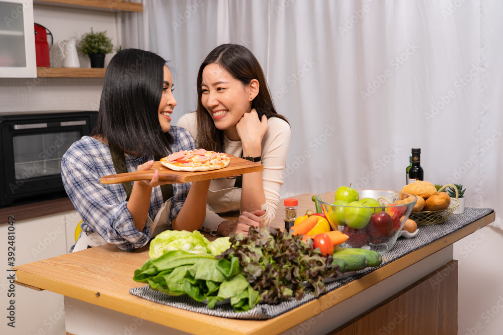 A happy young Asian lesbian couple standing behind wooden kitchen table with various vegetables and fruits, looking at each other with sweet smile, one holding a wooden tray with pizza.