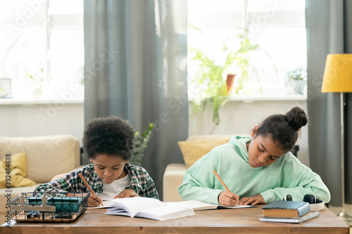 Cute diligent girl and boy in casualwear doing homework by table in living-room