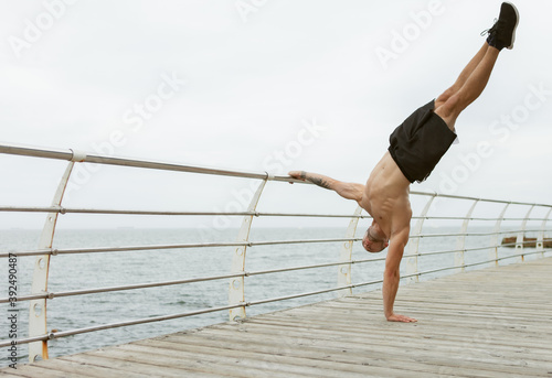 Muscular man athlete stands on his arms holding handrail on the beach