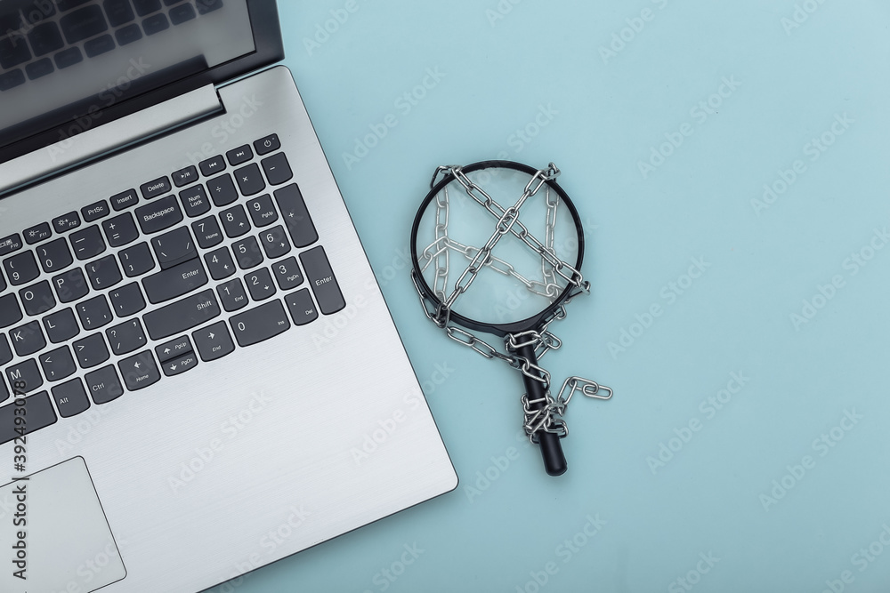 Laptop and Magnifying glass wrapped in a steel chain on blue background. Top view Laptop and Magnifying glass wrapped in a steel chain on blue background. Top view