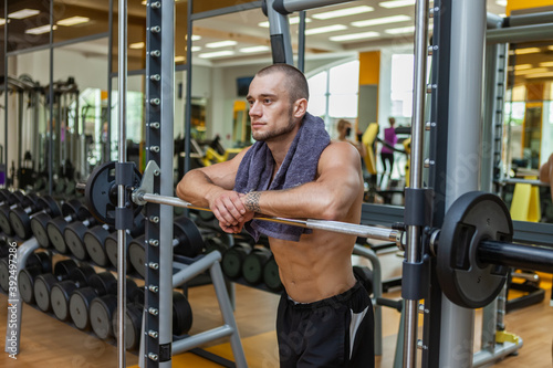 Tired muscular man with a naked torso and a towel on his neck resting on a barbell in the gym