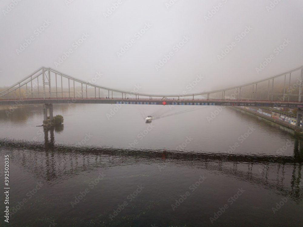 Fototapeta premium Aerial drone view. A motor boat sails under a footbridge on the Dnieper on a foggy morning.