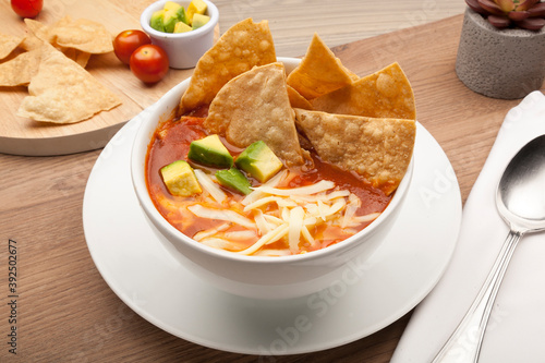 Mexican soup with tortilla chips, avocado and cheese in a white bowl on a rustic wooden table