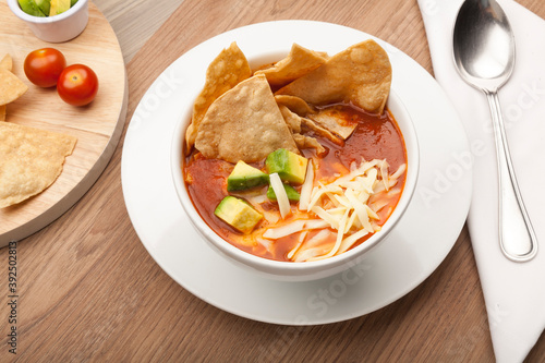 Mexican soup with tortilla chips, avocado and cheese in a white bowl on a rustic wooden table