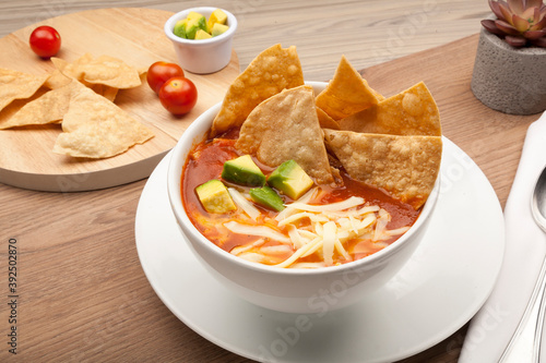Mexican soup with tortilla chips, avocado and cheese in a white bowl on a rustic wooden table