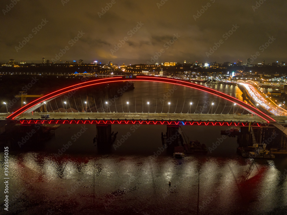 Obraz premium Aerial drone view. Modern arch cable-stayed bridge in Kiev in the evening. Colored lighting of the arch of the bridge.