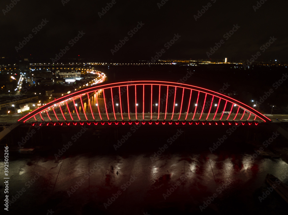 Obraz premium Aerial drone view. Modern arch cable-stayed bridge in Kiev in the evening. Colored lighting of the arch of the bridge.