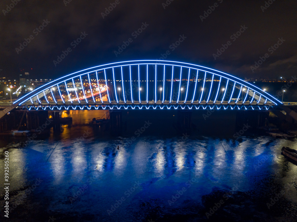 Fototapeta premium Aerial drone view. Modern arch cable-stayed bridge in Kiev in the evening. Colored lighting of the arch of the bridge.