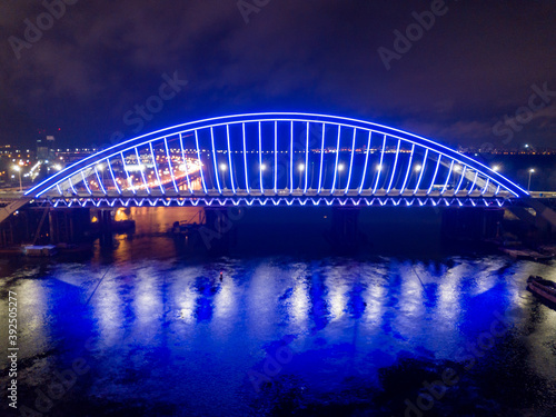 Aerial drone view. Modern arch cable-stayed bridge in Kiev in the evening. Colored lighting of the arch of the bridge.