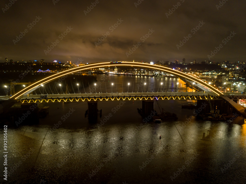 Naklejka premium Aerial drone view. Modern arch cable-stayed bridge in Kiev in the evening. Colored lighting of the arch of the bridge.