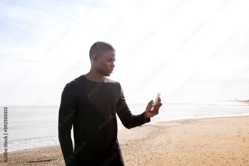 Black sportsman boy looks at his cell phone on the beach with a back light. Concept of sport and health. Black sportsman boy looks at his cell phone on the beach with a back light. Concept of sport and health.
