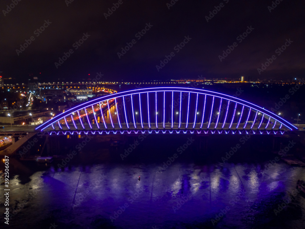Aerial drone view. Modern arch cable-stayed bridge in Kiev in the evening. Colored lighting of the arch of the bridge. Aerial drone view. Modern arch cable-stayed bridge in Kiev in the evening. Colored lighting of the arch of the bridge.