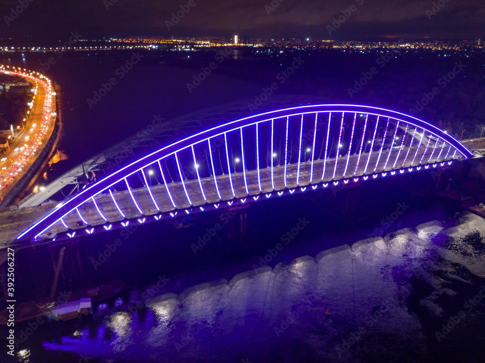 Obraz premium Aerial drone view. Modern arch cable-stayed bridge in Kiev in the evening. Colored lighting of the arch of the bridge.