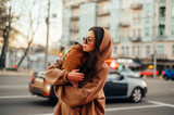 Stylish woman in casual clothes with a dog in her arms walks down the street, looking away. Lady carries in her arms a cute dog biewer terrier
