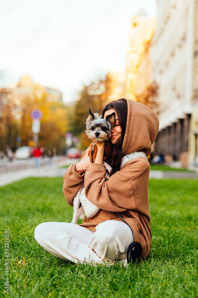 Beautiful woman sitting on the lawn with a cute little dog in her arms on a street background and playing with a pet. Vertical