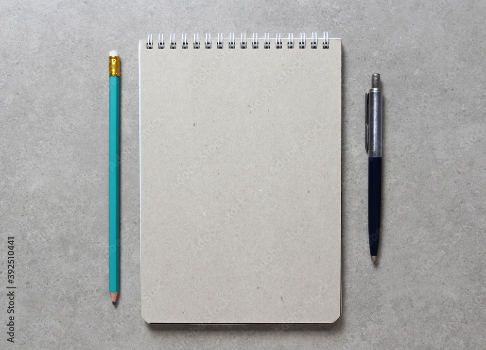 A spring notebook with a sheet of craft paper A5 with a ballpoint pen and simple green graphite pencil on light grey concrete background. Concept of new idea, business plan and strategy. A spring notebook with a sheet of craft paper A5 with a ballpoint pen and simple green graphite pencil on light grey concrete background. Concept of new idea, business plan and strategy.