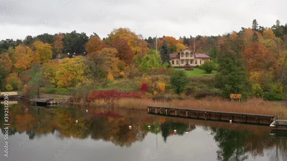 Mansion dream house at lake in autumn. Yellow orange and red plants and trees surrounding ocean front building. Old large home with dock or pier in fall. Forest behind and classy water on calm day Mansion dream house at lake in autumn. Yellow orange and red plants and trees surrounding ocean front building. Old large home with dock or pier in fall. Forest behind and classy water on calm day