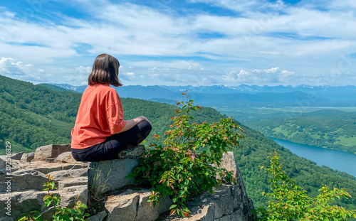 A woman with a bob hairstyle in a red T-shirt sits on the edge of a cliff