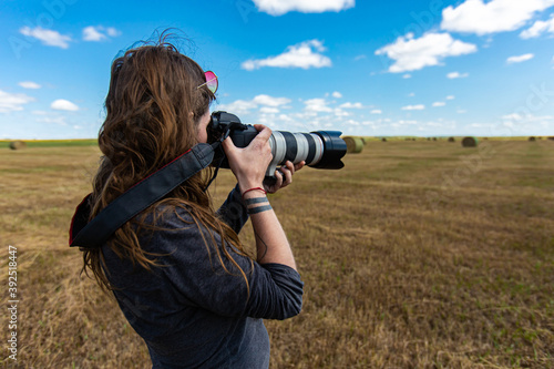 Young female photographer with long hair taking pictures outdoor with a telephoto zoom in a rural environment.