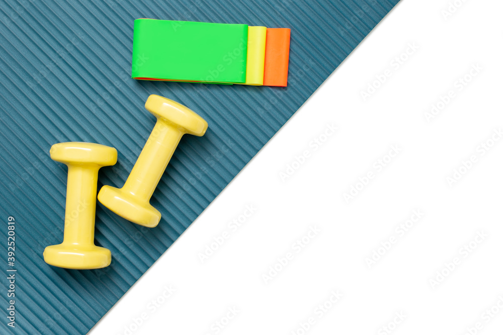 Yellow dumbbells and a set of multi-colored elastic bands for fitness on a sports mat and a white background diagonally. Yellow dumbbells and a set of multi-colored elastic bands for fitness on a sports mat and a white background diagonally.