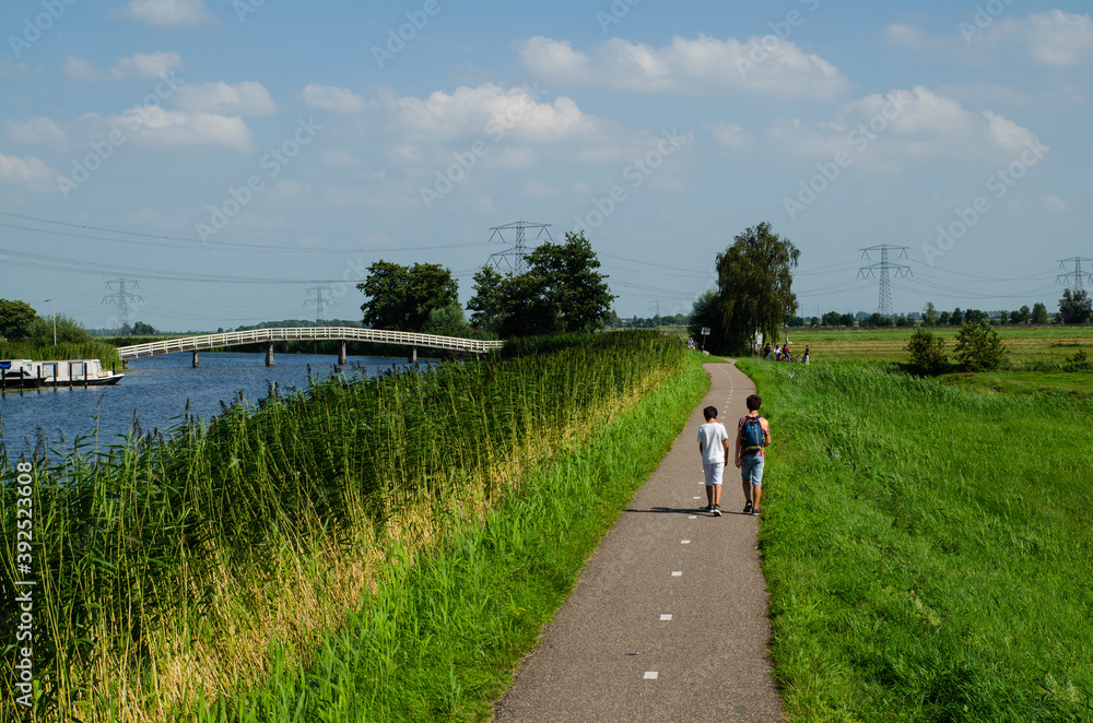 Obraz premium Kinderdijk, The Netherlands, August 2019. A small road winds through the Dutch countryside, lined with green grass and a canal, some boys are walking along it. On the left a white bridge.