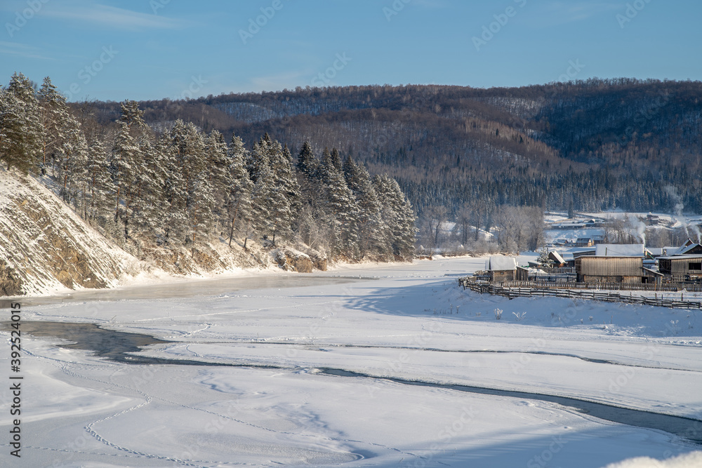 Winter, frosty day in a village located among the snow-capped mountains on the banks of the river. On the slopes of the mountains there is a coniferous forest.