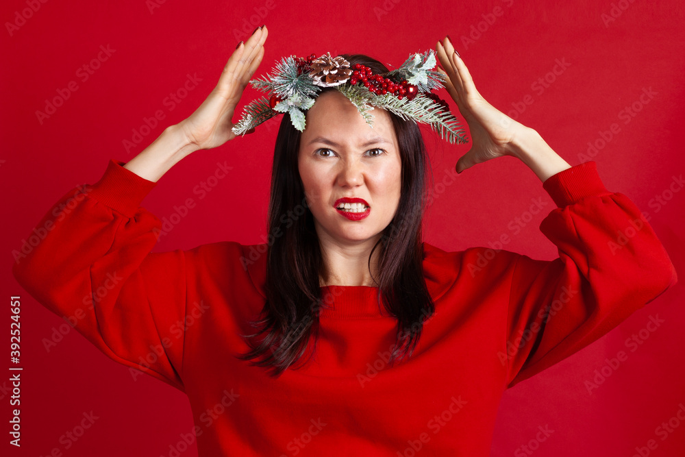 close-up portrait of a nervous, aggressive Asian young woman in a Christmas wreath with hands holding her head, on a red background. close-up portrait of a nervous, aggressive Asian young woman in a Christmas wreath with hands holding her head, on a red background.