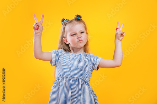 Funny little girl showing peace gesture on yellow background..