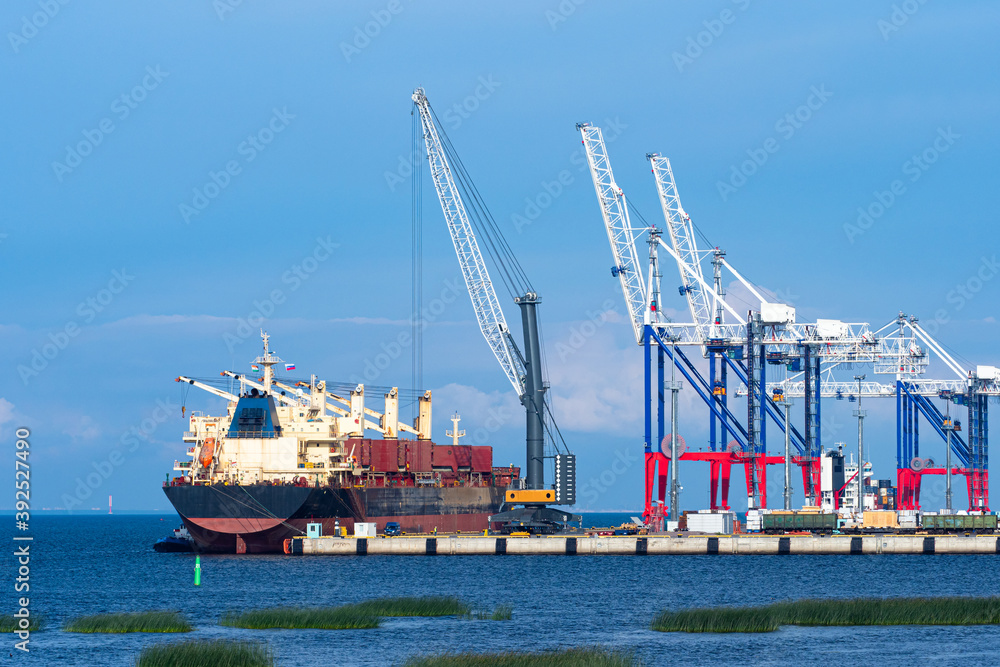 Cargo seaport. Terminal for loading sea containers. Loading cranes on shore of cargo port. Crane is loading something onto ship. Marine ship during shipment. Summer day. Unloading a sea vessel