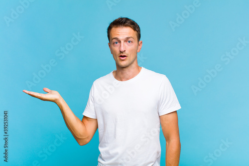 young handsome man looking surprised and shocked, with jaw dropped holding an object with an open hand on the side photo