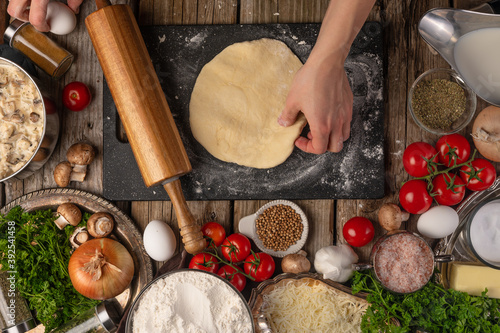 Chef hands with the dough and rolling pin on wooden table with variety of ingredients background. Concept of cooking process. Backstage of preparing tasty meal. View from above.